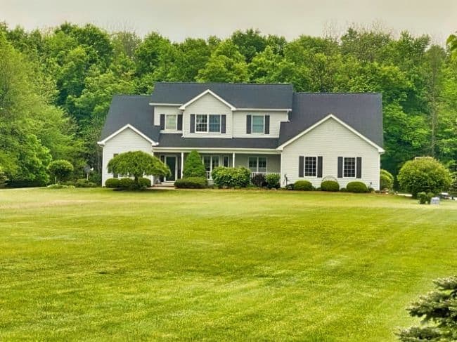 two-story home with white vinyl siding and gray shingled roof set back from the road