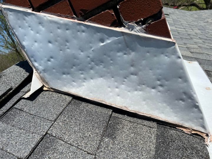 sideview of piece of silver metal flashing attached to a chimney on a shingle roof with dents to demonstrate what does hail damage look like on metal flashing